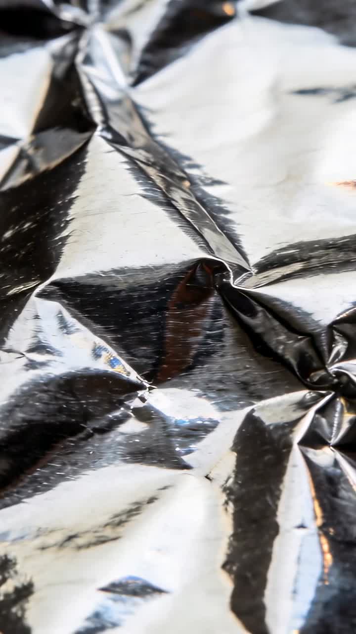 Video verticale: Telecamera in movimento che mostra un foglio di alluminio accartocciato con riflessi cangianti e piccole particelle in macro.
