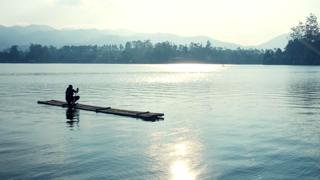 Man Paddles Small Raft On Shimmering Lake Surrounded By Mist Covered Hills