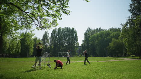 una familia juega al fútbol al aire libre, un niño ata su cordón de los zapatos cerca de un poste de la puerta mientras otro atrapa una pelota, otros juegan cerca, rodeados de vegetación y un cielo despejado