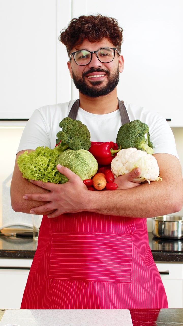 Giovane uomo arabo sorridente che tiene verdure fresche nella cucina di casa per la preparazione di pasti sani per la dieta.