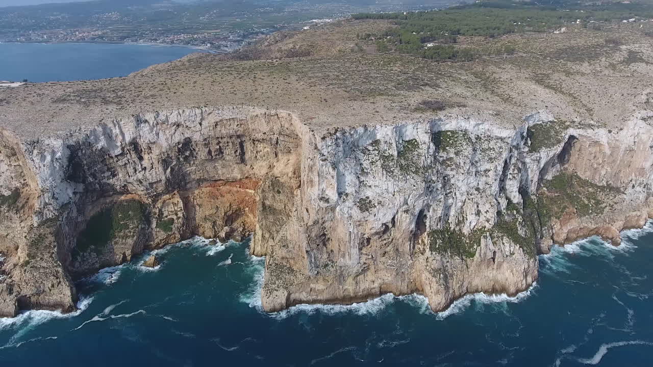 Aerial view flying backwards of Cape San Antonio, Denia, Valencia. View of Rocky cliff and sea waves crushing on the coastline. Blue water in a sunny day