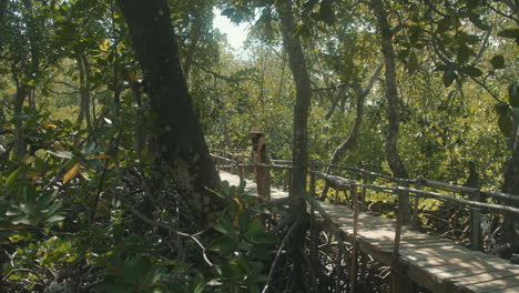 General-shot-of-a-Caucasian-woman-wearing-a-straw-hat-on-a-wooden-bridge-looking-at-and-enjoying-the-green-forest-around-her