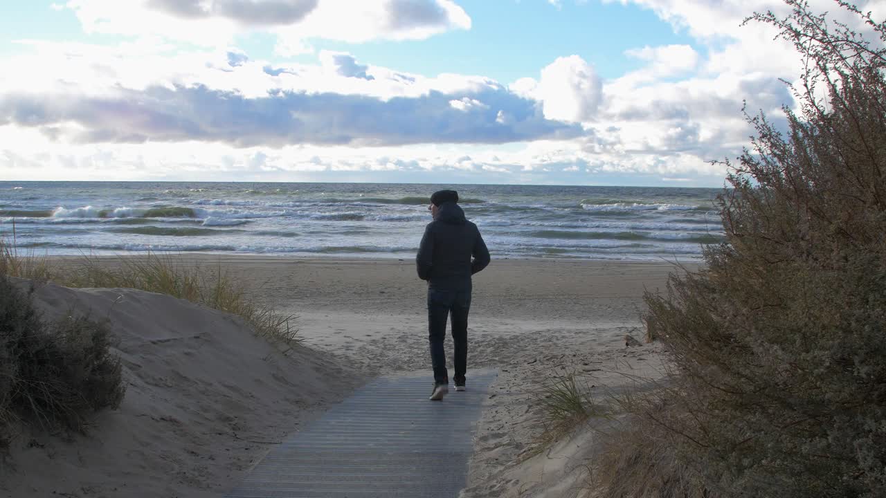 vista posterior de un hombre caucásico explorando el bosque costero nórdico, un camino de madera, un hombre caminando solo hacia la playa de arena blanca del mar báltico, un día soleado con nubes, un concepto de actividad saludable, una toma amplia