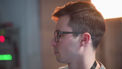 person working in tv studio, focused on monitor displaying broadcast setup. close-up of face with glasses as individual adjusts video equipment, studio lights in background
