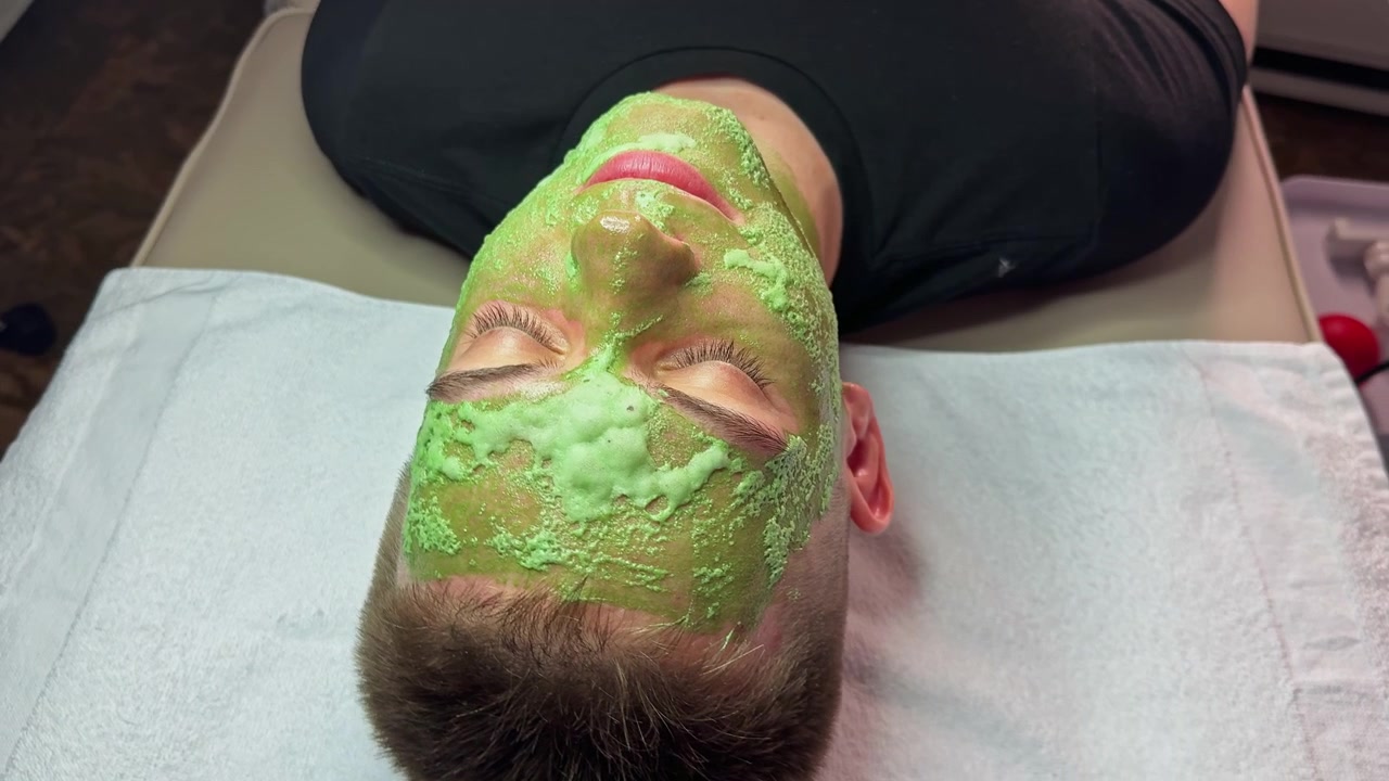 Boy receiving a facial treatment at a spa, with green mask applied, relaxing on a treatment table in a wellness center during daytime hours