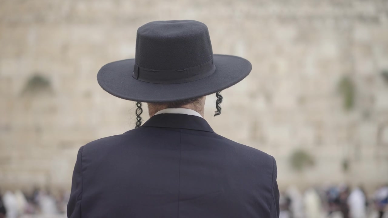 Jüdischer Mann betet an der Klagemauer in Jerusalem, Israel. Er hat lange Schläfenlocken und trägt einen schwarzen Hut und Mantel. Die Kamera fährt in Zeitlupe mit selektivem Fokus auf das Motiv zu, von hinten gesehen.