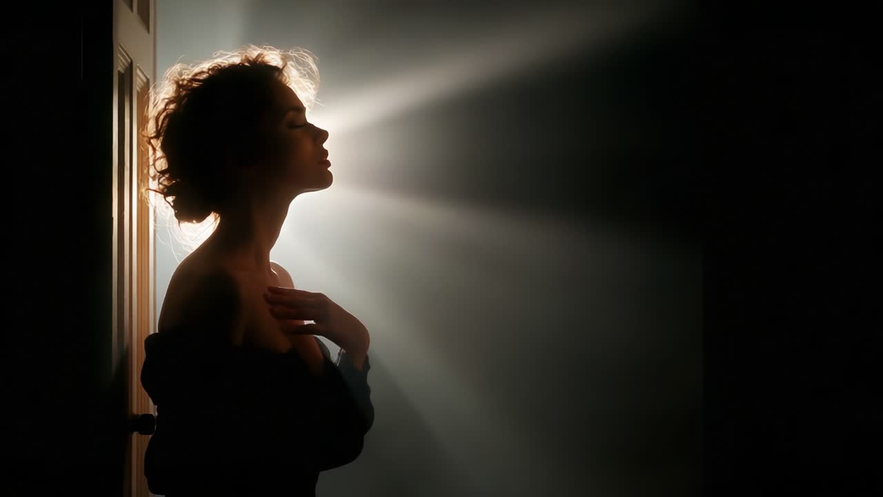 A woman stands in silhouette against a backdrop of radiant beams of light, evoking a sense of mystery and elegance as shadows play around her serene form in a dimly lit room