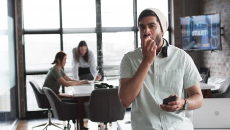 Young-Asian-business-man-with-headphones-around-his-neck-eats-an-apple,-holding-a-phone