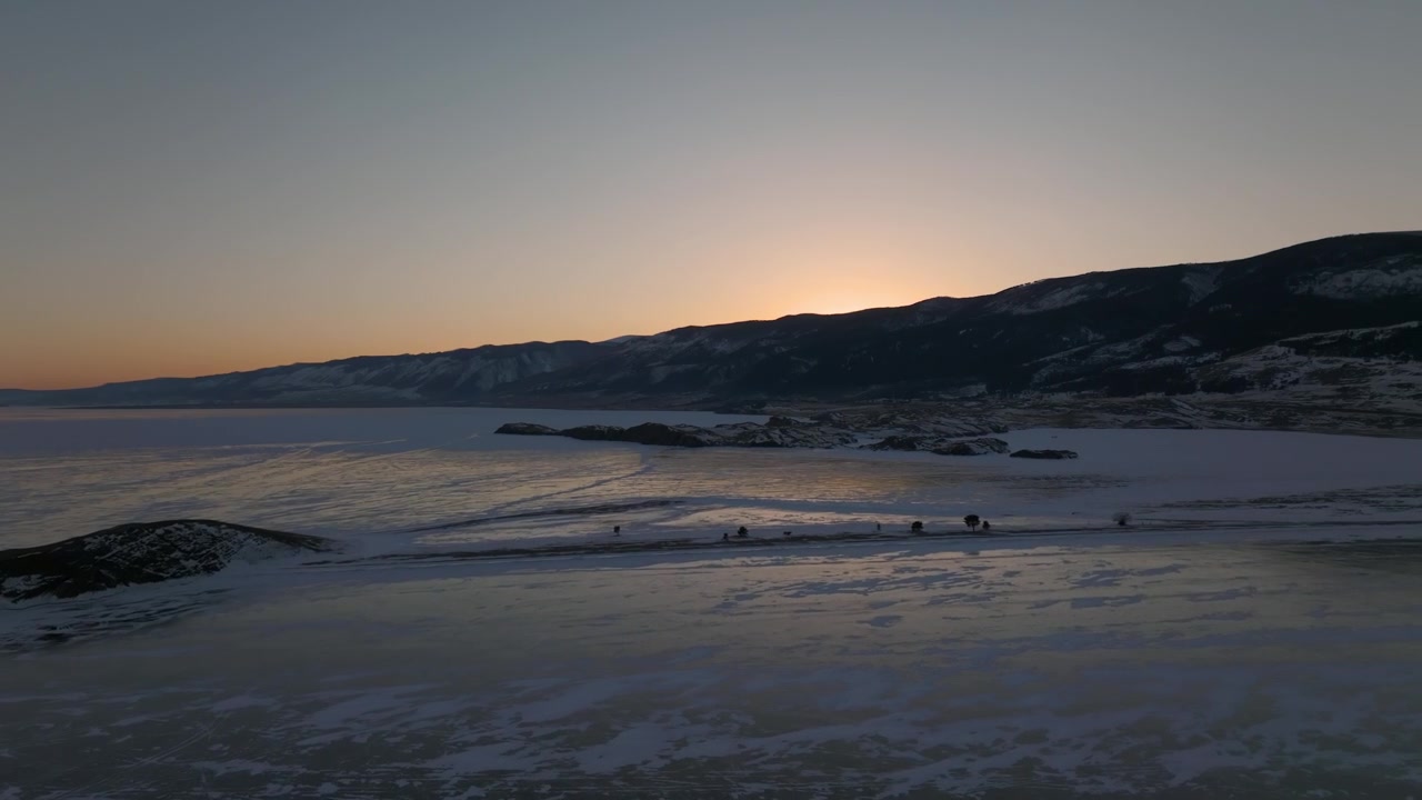 Luftoptagelse af den frosne Bajkalsø ved solnedgang, med revnet is, sneklædt kystlinje og mørk bjergryg i horisonten, et roligt vinterlandskab i Rusland.