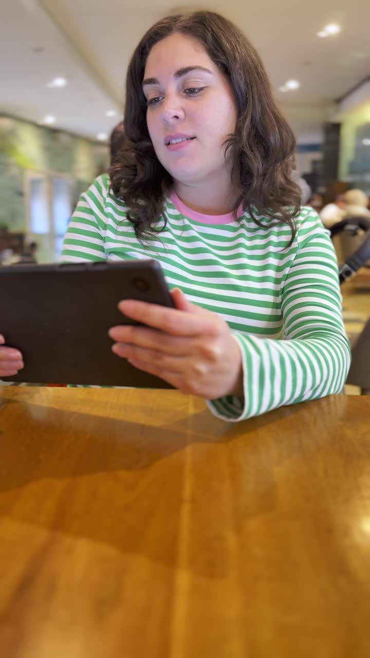 A woman is sitting at a table with a tablet in her hand, in coffee shop.