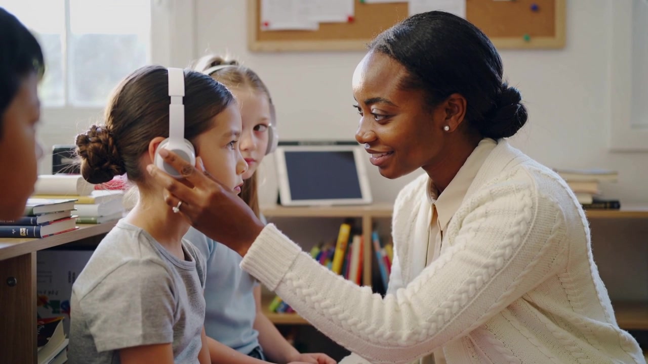 Placing teacher kneeling in cardigan adjusting white headset on student at school, preparing lesson