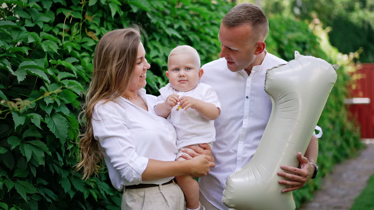 Loving Caucasian parents hold their baby kissing him on the head. Man is holding a balloon shaped like number one. Greenery at backdrop.