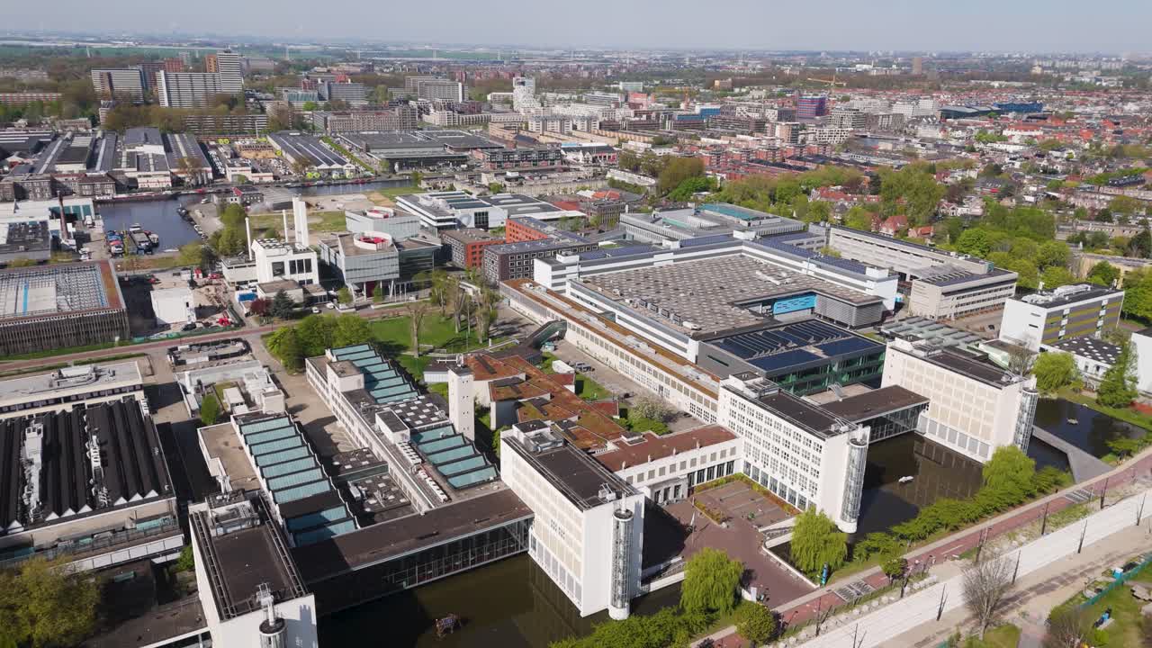 Drone footage of TU Delft’s science campus, highlighting research buildings, water features, and surrounding infrastructure in a sunny urban landscape.