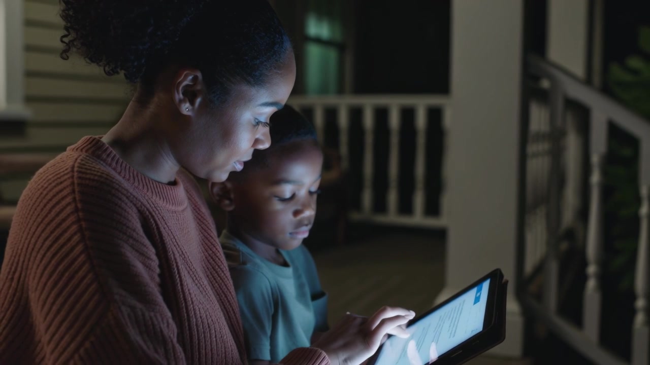 Leaning mother in rust sweater guiding son tapping tablet on porch at night, sharing screen glow