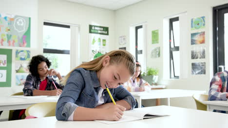 multiracial students in classroom smiling and writing in notebooks during school lesson