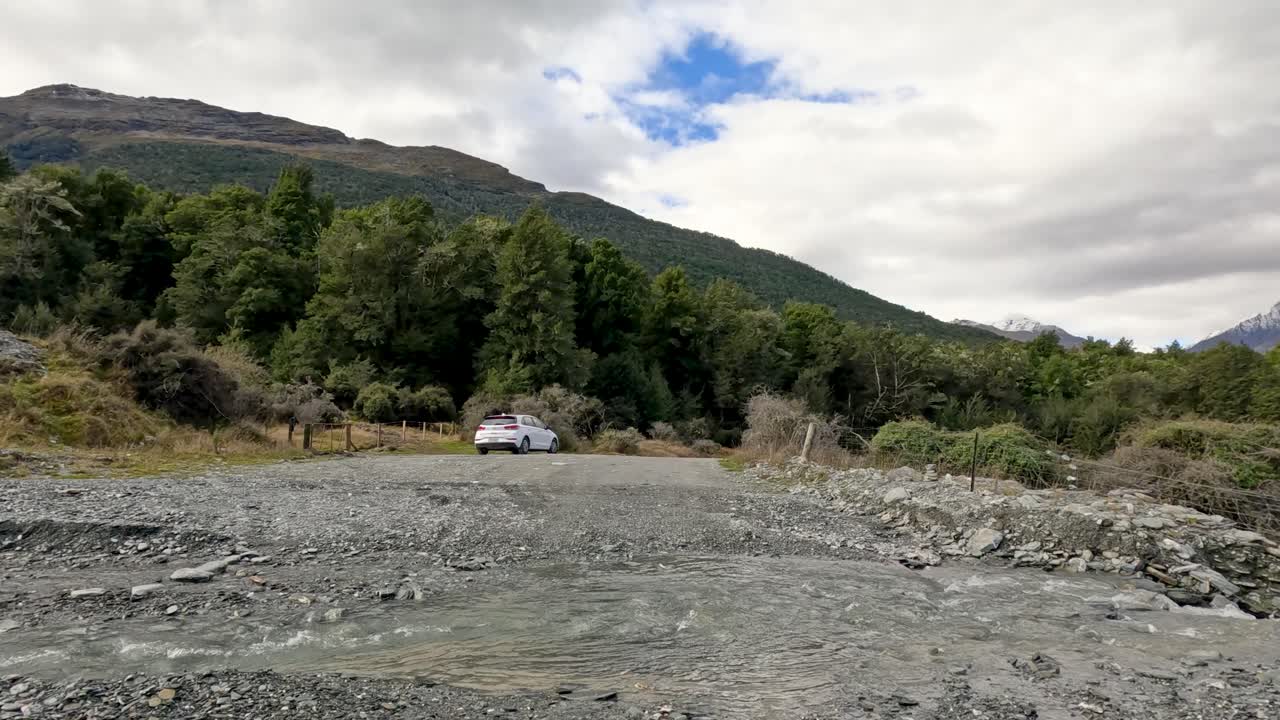 Vehicle approaches stationary car across stony riverbed, surrounded by forested hills under cloudy daylight