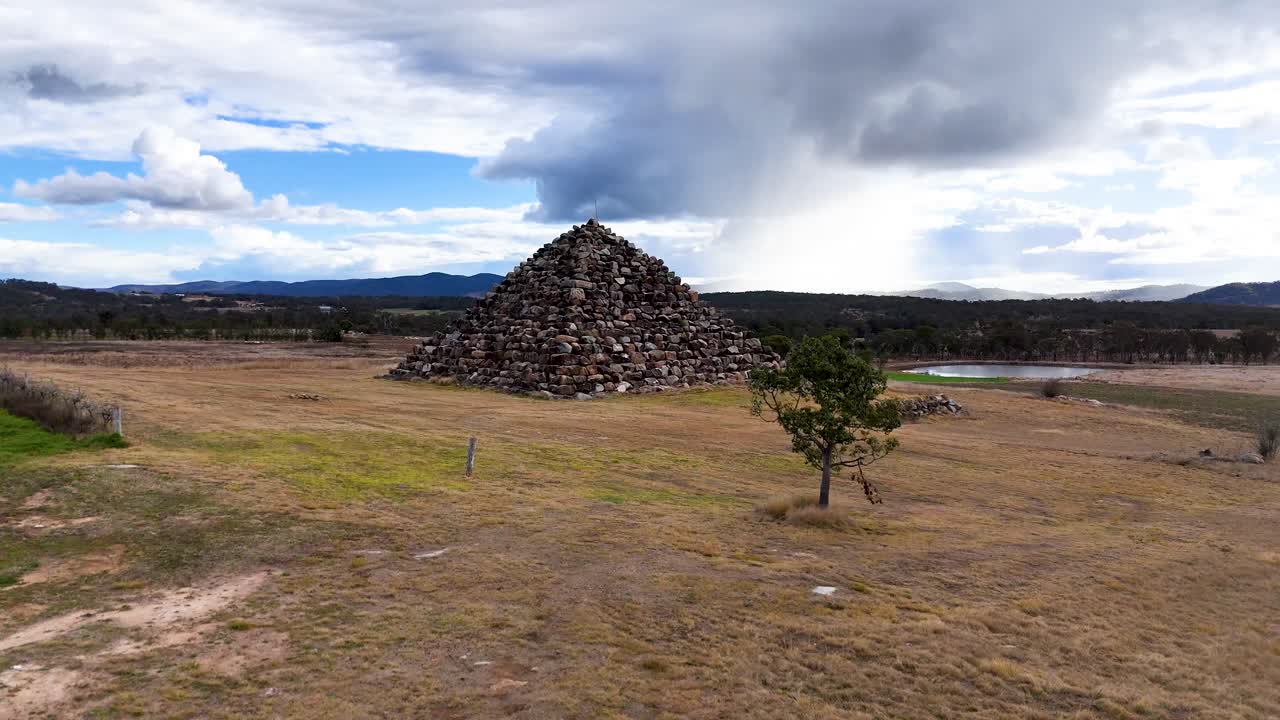 Drone footage circles a large stone pyramid structure in a dry, open field under dramatic skies near Stanthorpe, Queensland, with shifting light and perspective