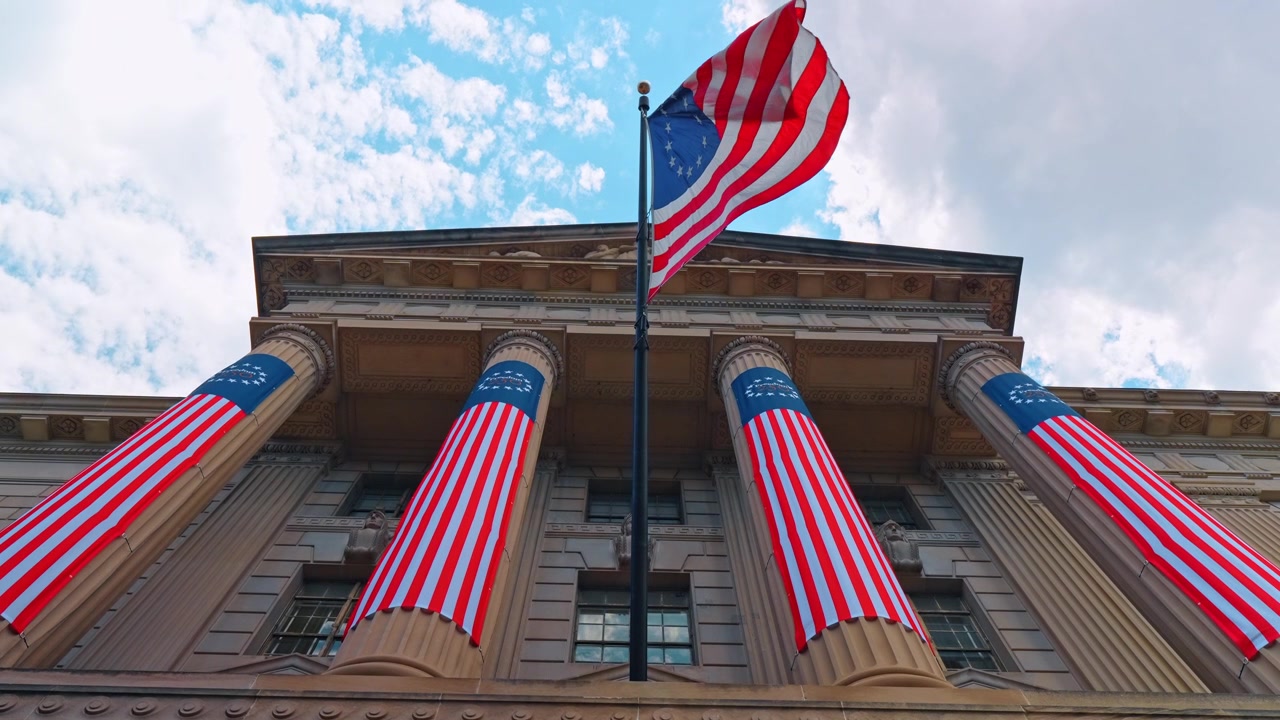 Bandiera degli Stati Uniti sopra le colonne di un edificio neoclassico, striscioni in tessuto coprono quattro colonne sotto le nuvole.