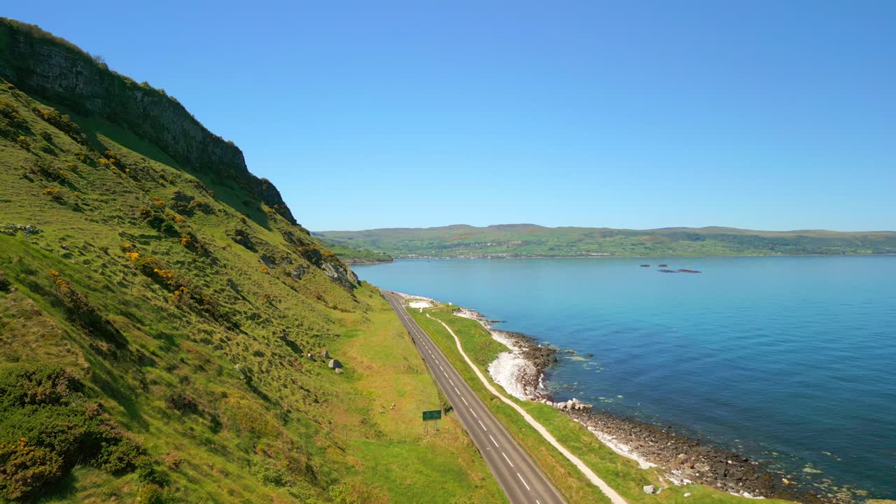 Advancing aerial video of the Causeway Coastal Route in County Antrim, Northern Ireland, UK, on a bright and sunny day. A road snakes along the cliffside. Filmed in 4K, 60FPS and with Rec709 color.