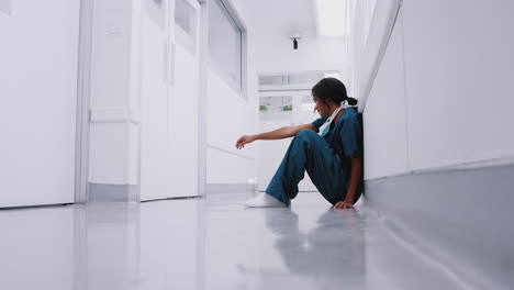 stressed and overworked female doctor wearing scrubs sitting on floor in hospital corridor