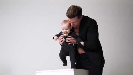 male wearing black suit holding a little child learning to walk. father and son concept. white backdrop.