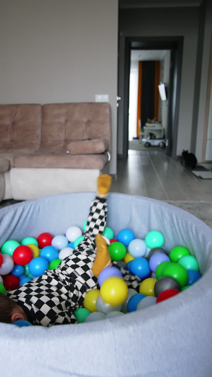 Energetic Caucasian toddler wearing checkered pajama approaches the dry pool. Happy kid jumps on the balls and moves them to the sides.