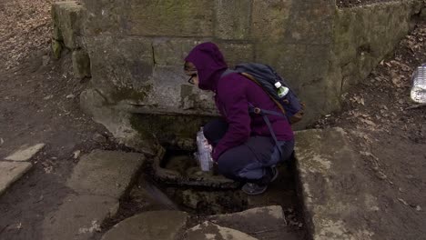 Frau-Auf-Einer-Wanderung,-Die-Ihre-Trinkflasche-Mit-Kaltem,-Klarem-Wasser-Aus-Einer-Quelle-Im-Wald-Füllt