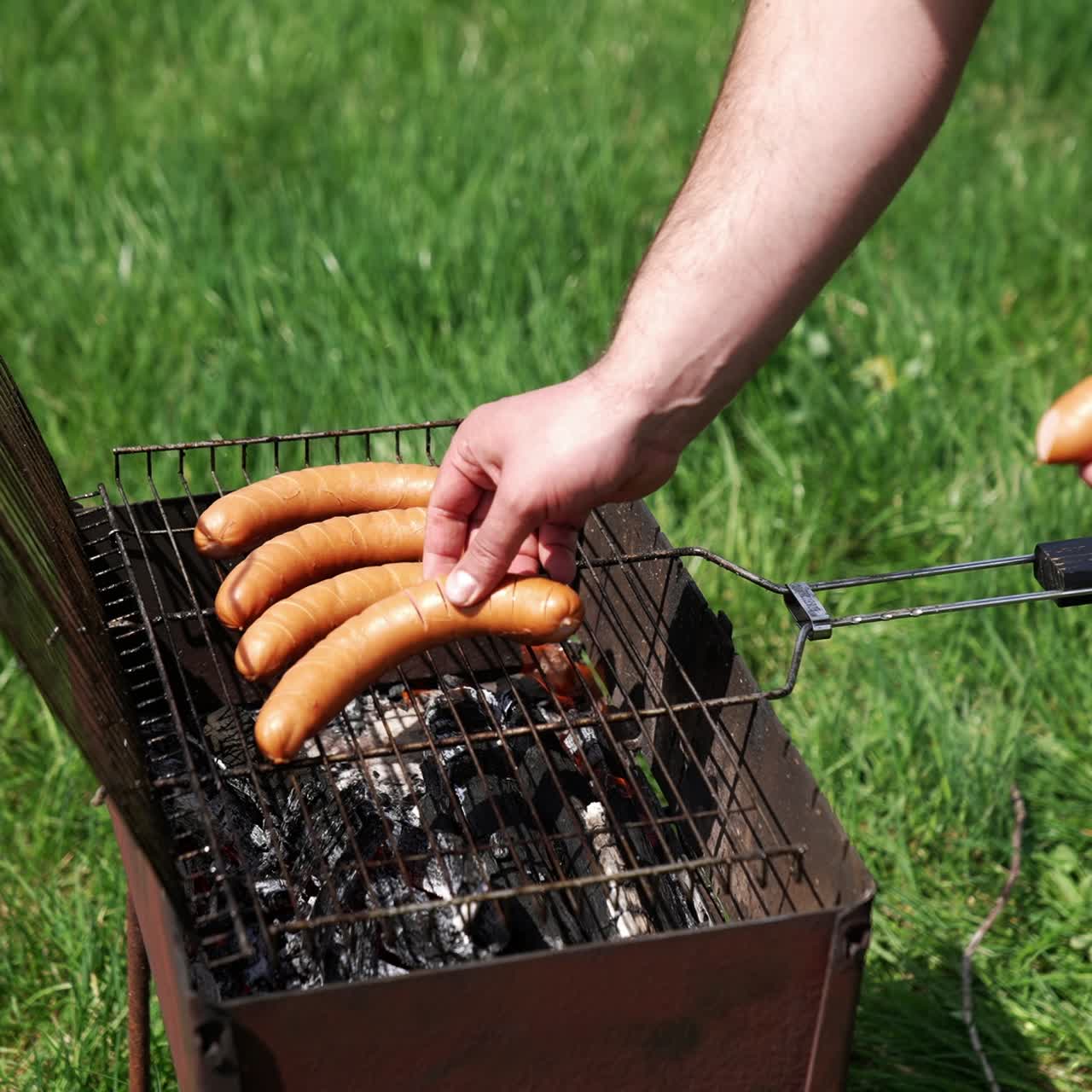 Man puts sausages into a grill for frying. Cooking hot dog sausages on fire. Barbecue in nature. Brazier on green grass background