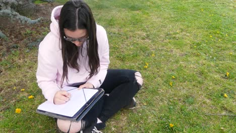 young teenage female writer with glasses writing on a paper sheet using a pen, sitting on grass outside