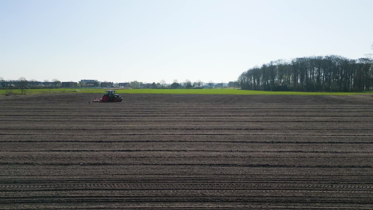 Wide shot of a red tractor plowing a large flat field in the Dutch countryside, symbolizing agriculture, farming, and springtime preparation.