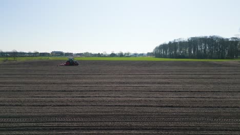 wide shot of a red tractor plowing a large flat field in the dutch countryside, symbolizing agriculture, farming, and springtime preparation.