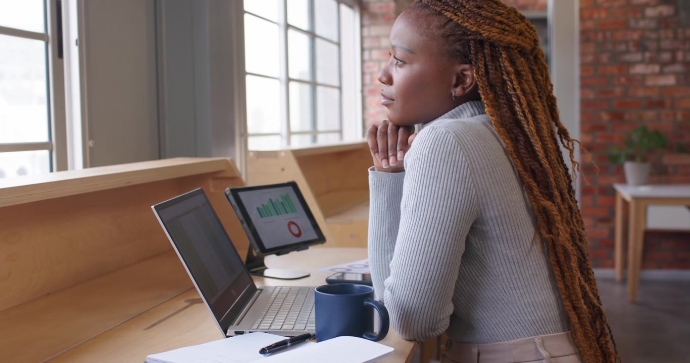 Donna africana di mezza età che alza lo sguardo dal laptop, con la mano sul mento, riflettendo su un grafico su tablet al bancone.