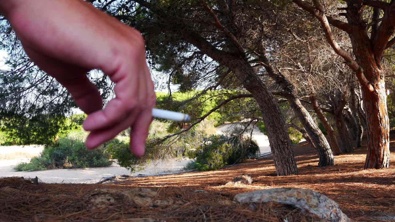 disparo cerrado de una mano con un cigarrillo frente a un bosque de pinos