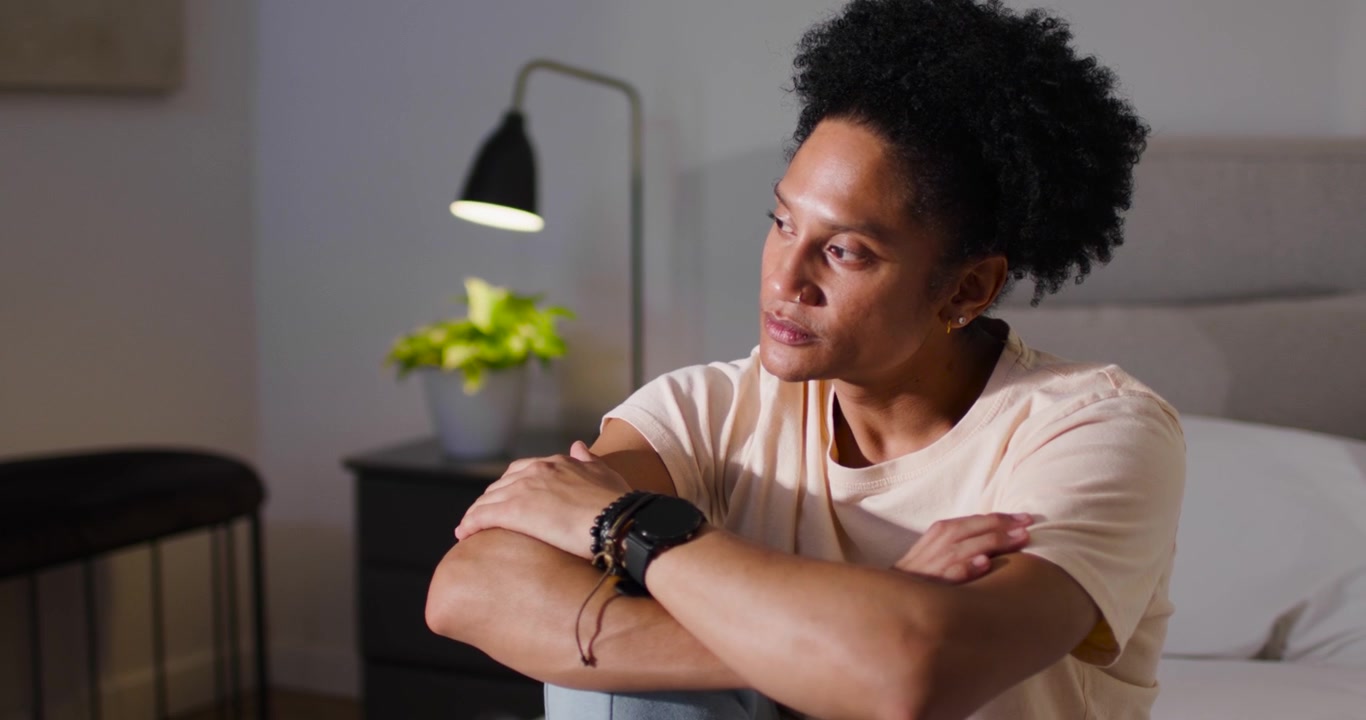 African American man sitting on bed hugging knee, turning gaze right, reflecting as lamp glows