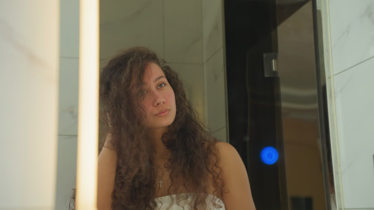 Una donna si abbellisce. Un'ospite sicura di sé acconcia i riccioli mentre ispeziona attentamente il suo riflesso. Una donna avvolta in un asciugamano prova pose e sistema i gioielli nello specchio del bagno dell'hotel, illuminato caldamente.