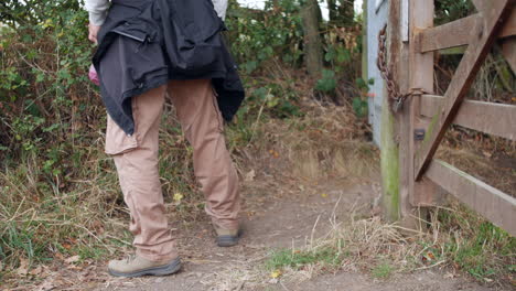 Ein-Mann-Mit-Einem-Spazierstock-Auf-Einer-Wanderung,-Der-Ein-Tor-öffnet-Und-Durchgeht,-Bevor-Er-Es-Schließt,-Während-Er-Durch-Die-Landschaft-Wandert