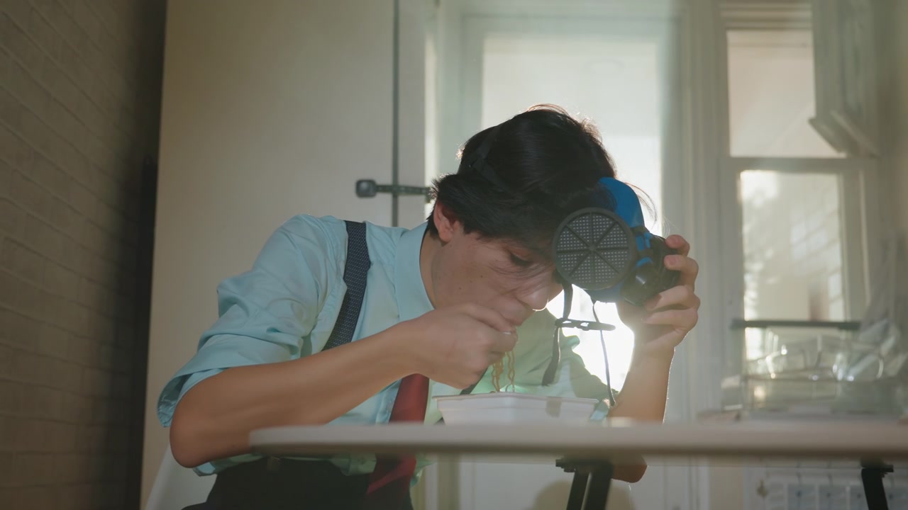 窓際で青いマスクをテストするアジア人男性、シャツとサスペンダーを着用した若いアジア人メーカーが呼吸器のような装置を検査する、強い逆光がドラマチックなレンズフレアを生み出し、集中してストラップを調整する手元