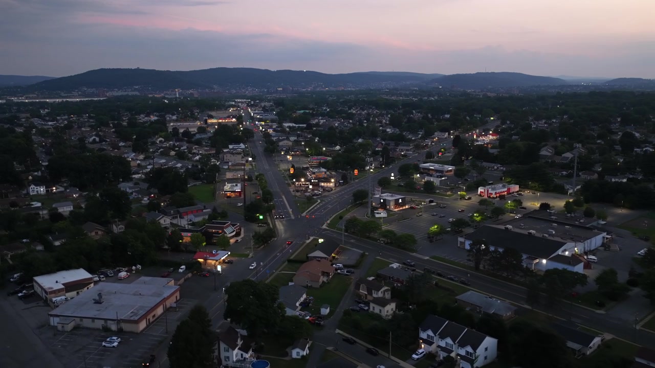 L'incrocio di una piccola città risplende di insegne illuminate e lampioni, case e alberi fitti si estendono verso colline distanti al crepuscolo nella Lehigh Valley, Pennsylvania, USA. Ampia inquadratura aerea con drone.