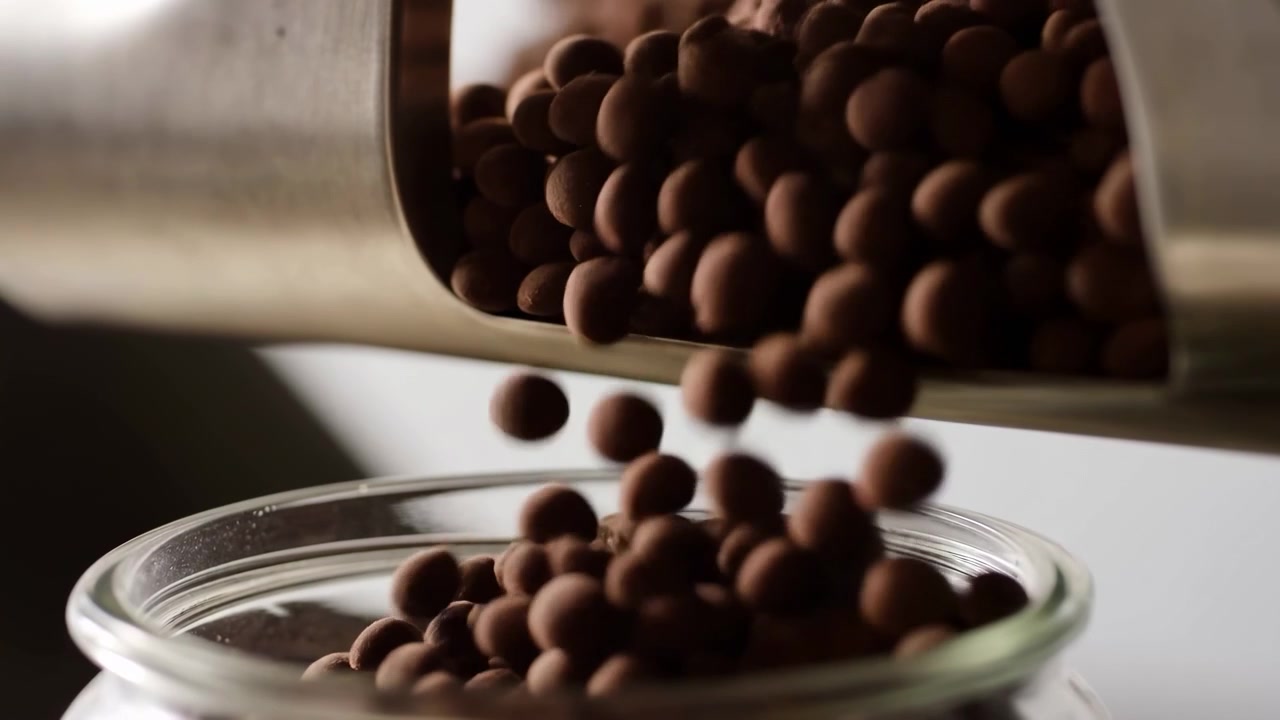 Caramelle sferiche ricoperte di cioccolato cadono da un cucchiaio di acciaio inossidabile in un barattolo di vetro trasparente, pezzi catturati a mezz'aria e che si accumulano all'interno del bordo con luce calda da studio, ombre morbide, ricca consistenza dolce