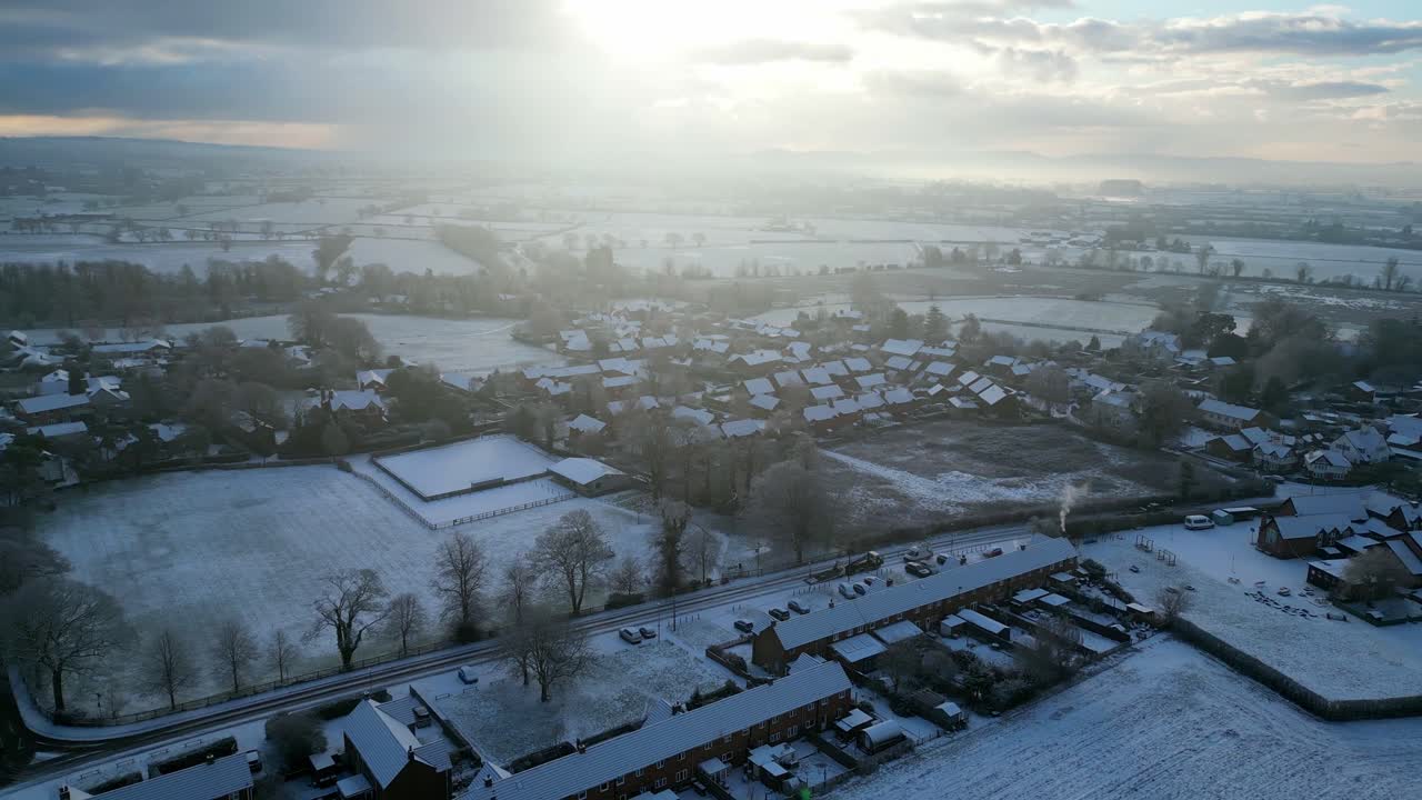 Aerial view of a quaint English village on a crisp winter morning, with snow-covered rooftops. The bright sunlight bathes the village in a golden glow, creating a serene and picturesque winter scene.