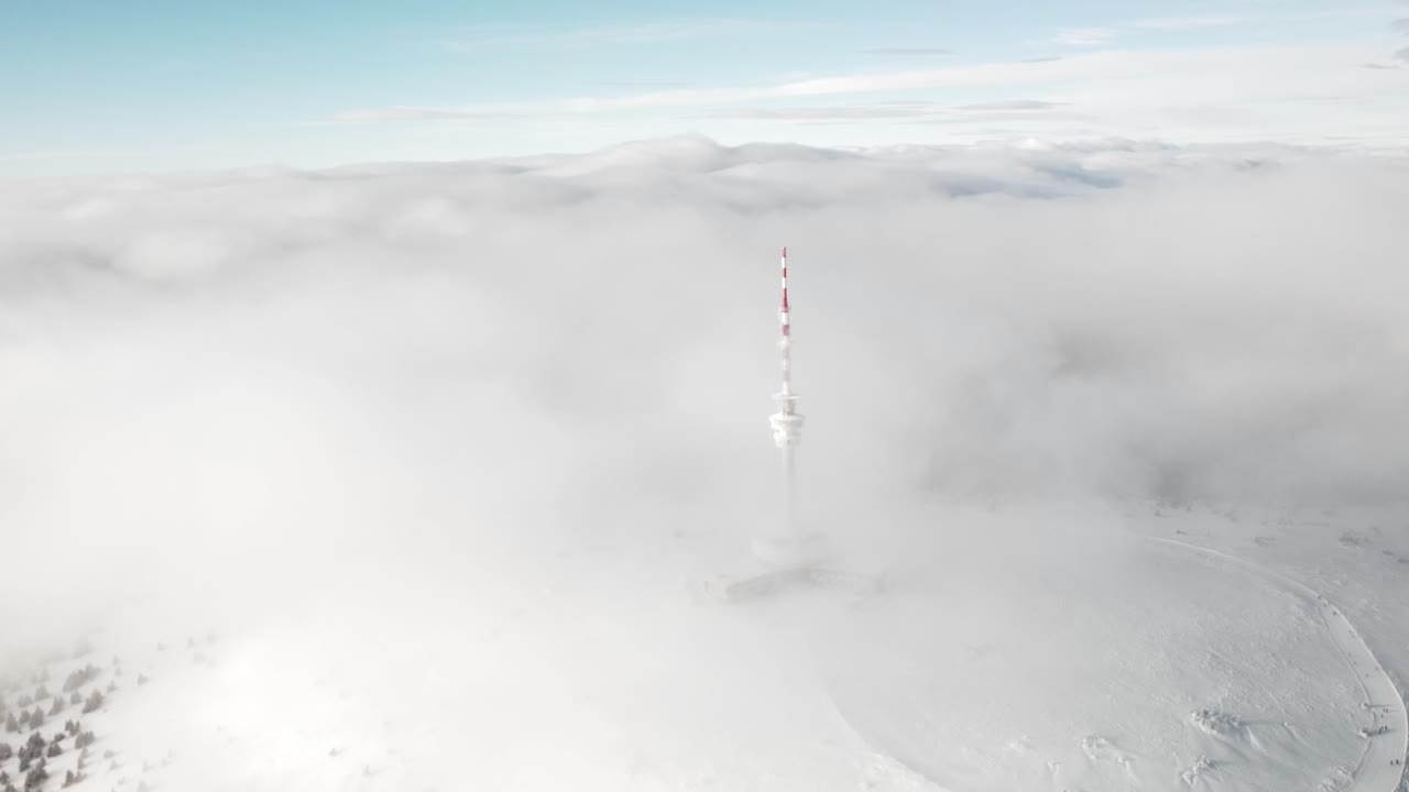 Drone slow approach to Praded iconic tower atop Jeseník mountains, framed by mist and fog in daylight, transmitter and receiver tower above hotel