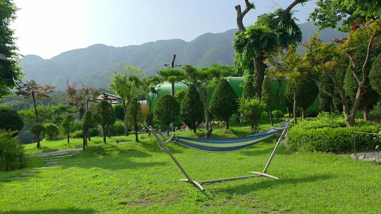 A striped hammock stretches across a vibrant green lawn surrounded by manicured trees and garden beds at Glamtree Resort in Gapyeong, South Korea, with mountains in the background