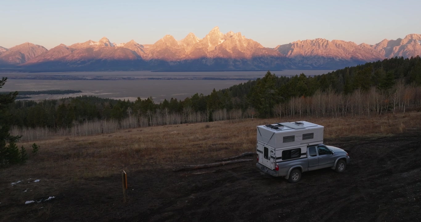 RV camper parked in field with Grand Teton mountains glowing at sunrise, aerial telephoto slow ascend
