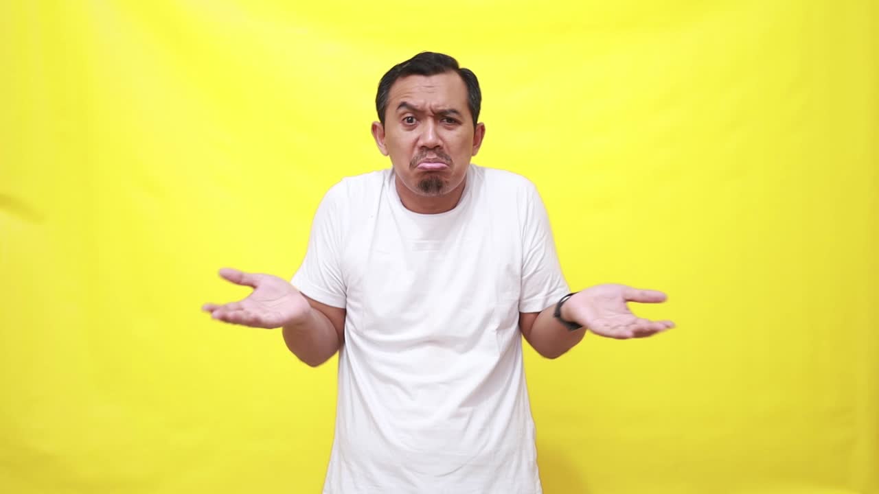 아시안 남자가 어깨를 끄덕여 무의식적으로 손을 들고, 아무런 단서도 없고, 노란색 배경에 대하여 포즈를 취할 수 없습니다.