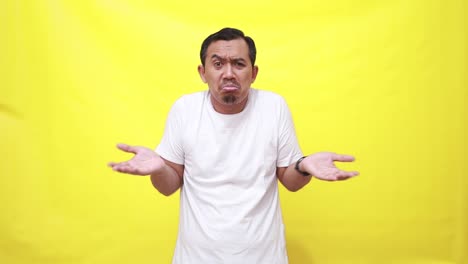 asian man shrugs shoulders unaware raises hand clueless has no idea cannot answer poses against yellow background