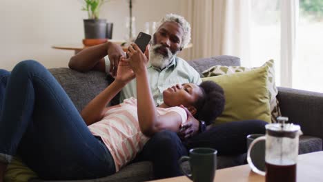 Happy-african-american-couple-relaxing-on-couch-drinking-coffee-and-looking-at-smartphone