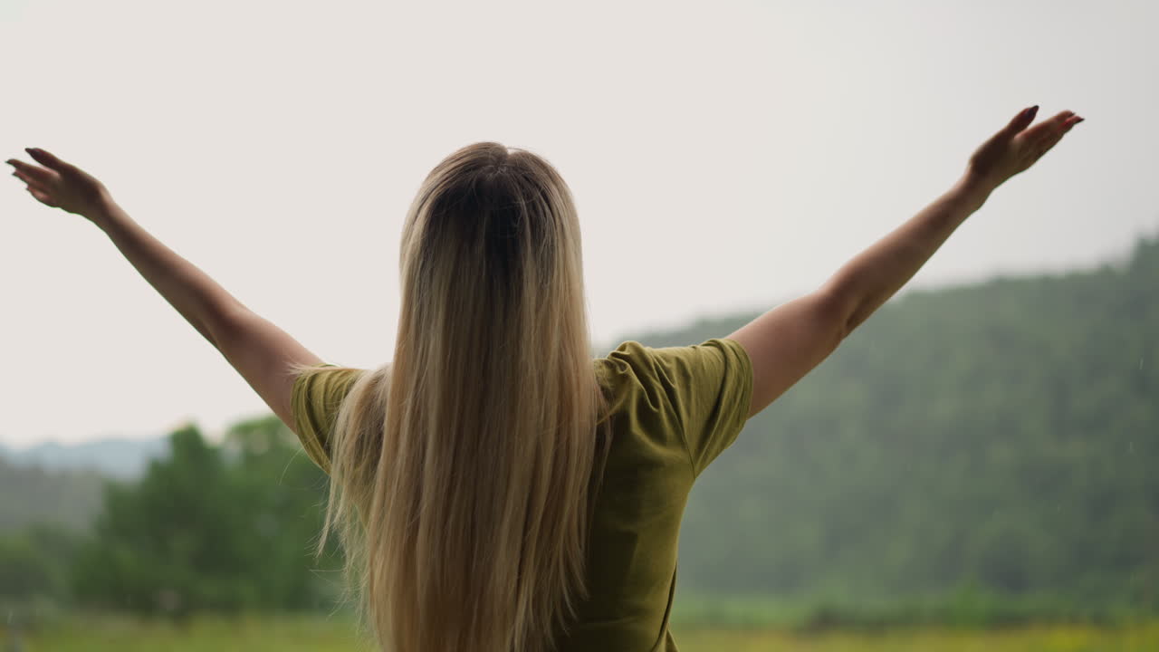 mujer levanta las manos feliz de la lluvia cálida contra la colina verde