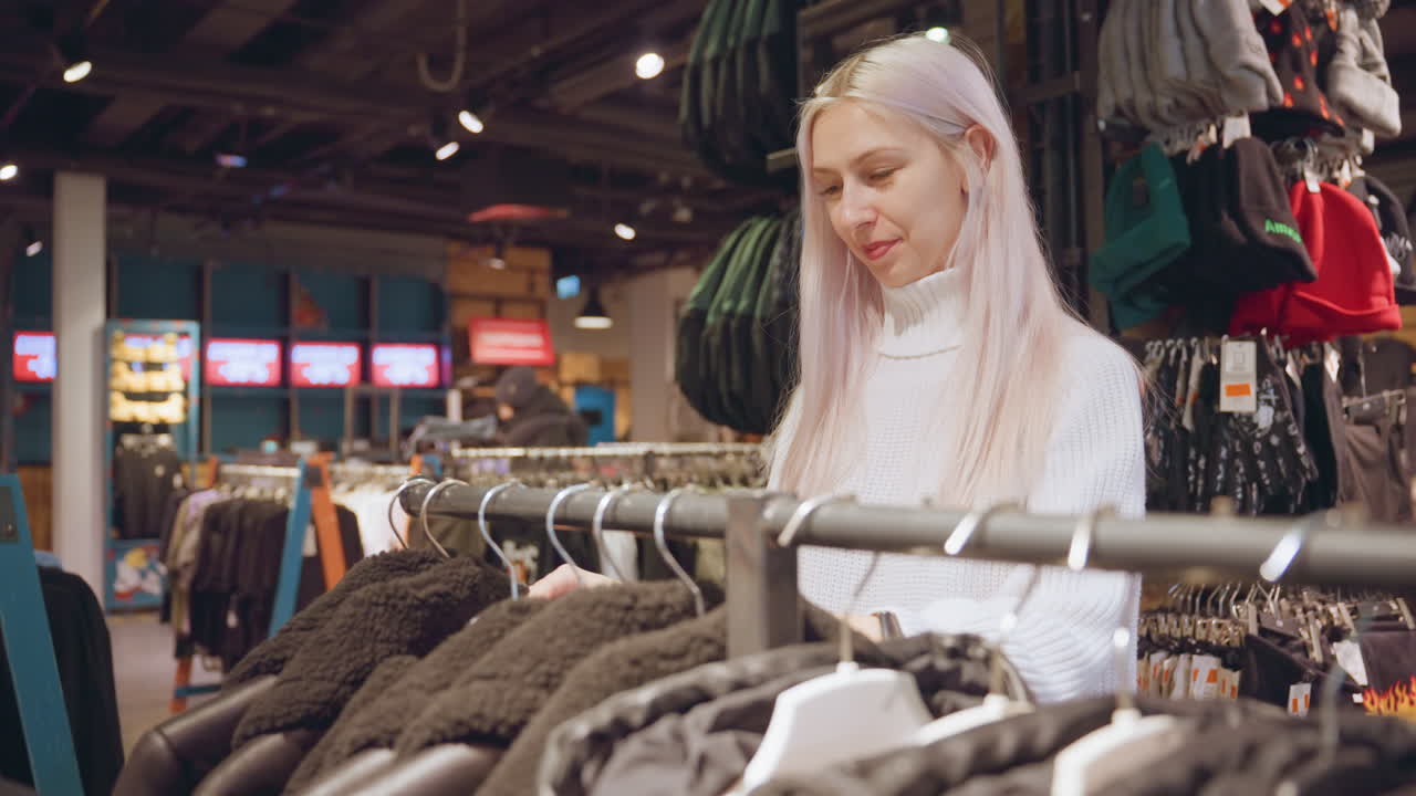 Smiling customer with silver hair in turtleneck explores jacket rack inside clothing store, gliding fingers over shearling coats while envisioning winter wardrobe upgrade under warm boutique lighting