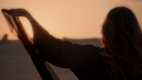 A-girl-holding-her-black-scarf-in-her-hands-and-waving-towards-the-camera-in-the-middle-of-the-Arabian-desert-wearing-an-abaya-while-sunset-close-up-shot-fossil-dunes