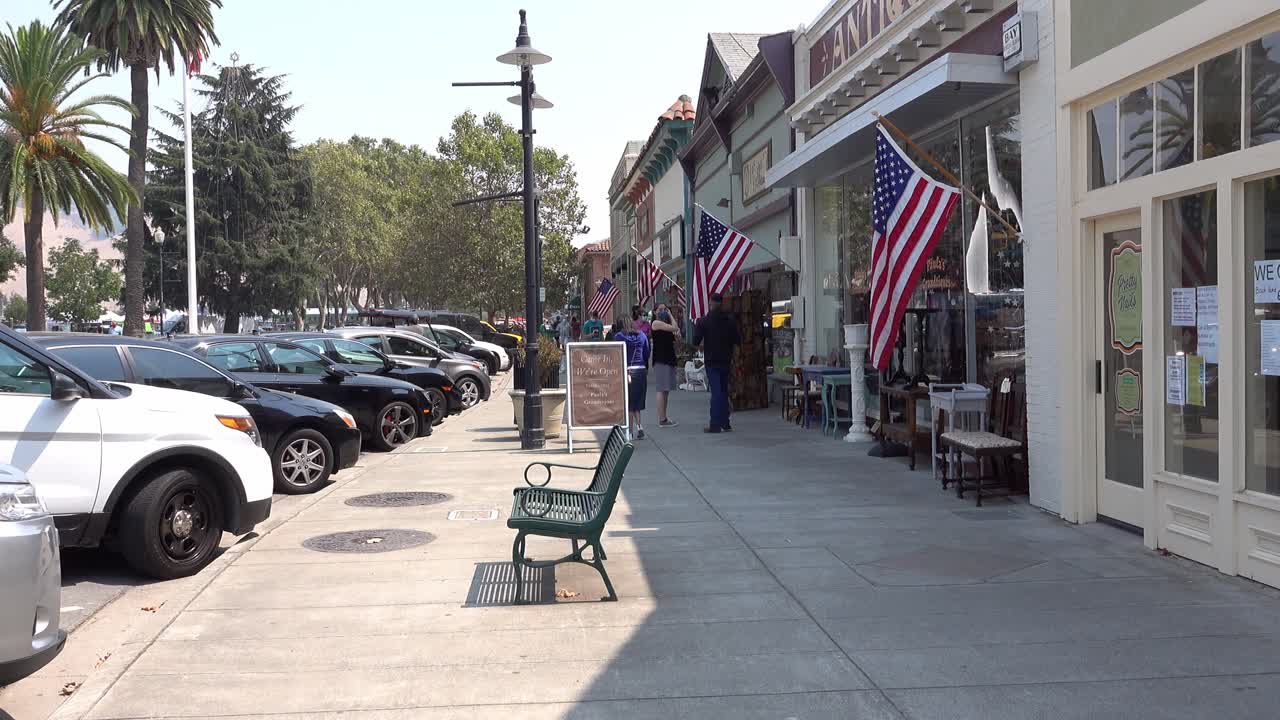 Camera pulling back on small town main street sidewalk bright summer's day with american flags flying